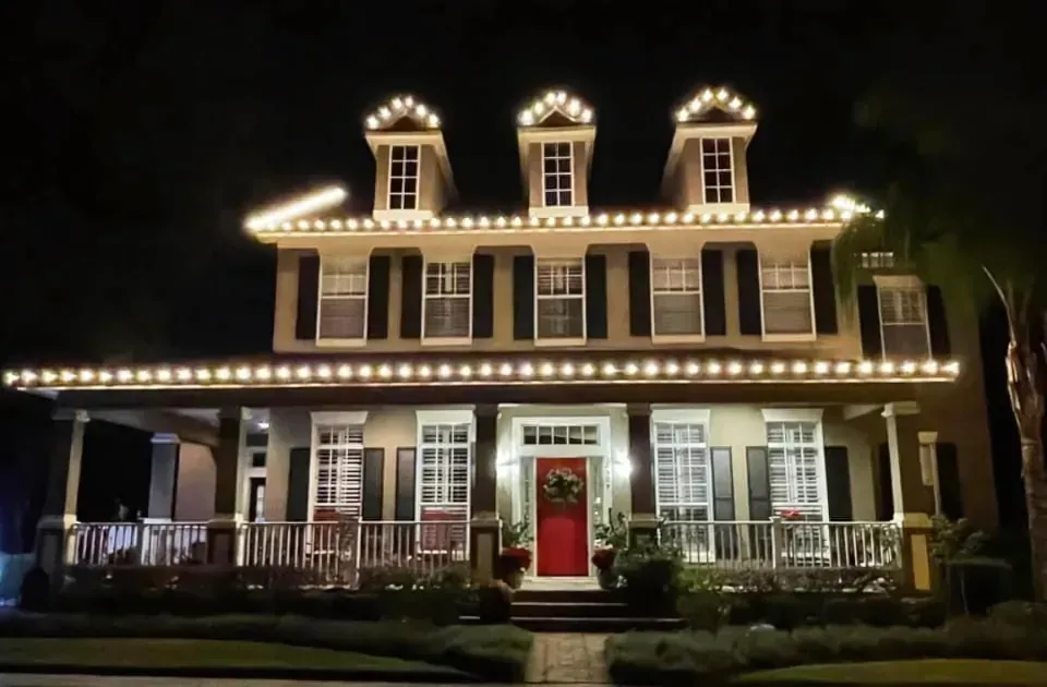 House decorated with white lights for Christmas at night. Red front door and green wreath.