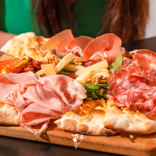 Assortimento di affettati e formaggi su una pizza tostata, servita su un tagliere di legno, con una persona sullo sfondo