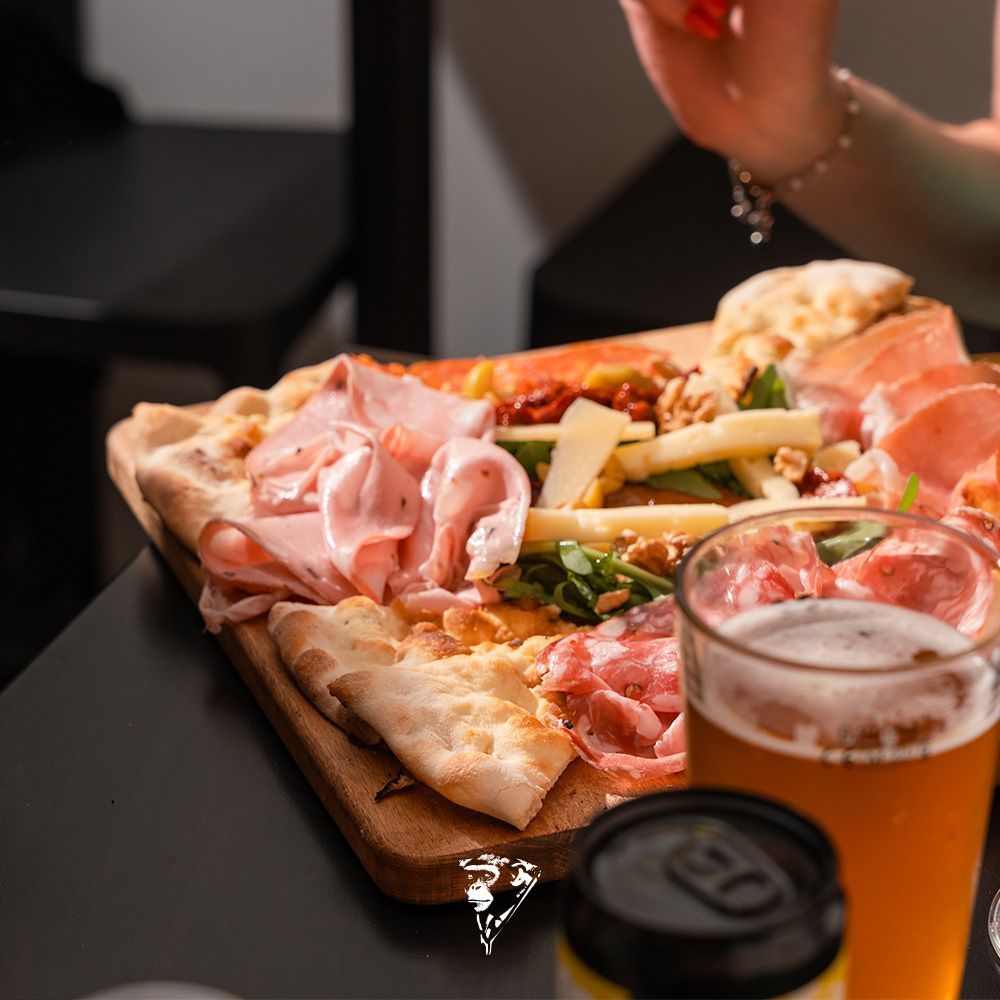 Tagliere di salumi e pizza con birra sul tavolo, con una mano che allunga la mano per prendere del cibo
