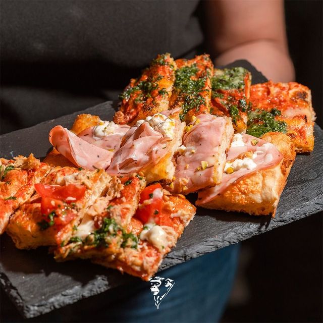 Una pizza quadrata tagliata a fette, condita con prosciutto ed erbe aromatiche, è adagiata su un vassoio di ardesia