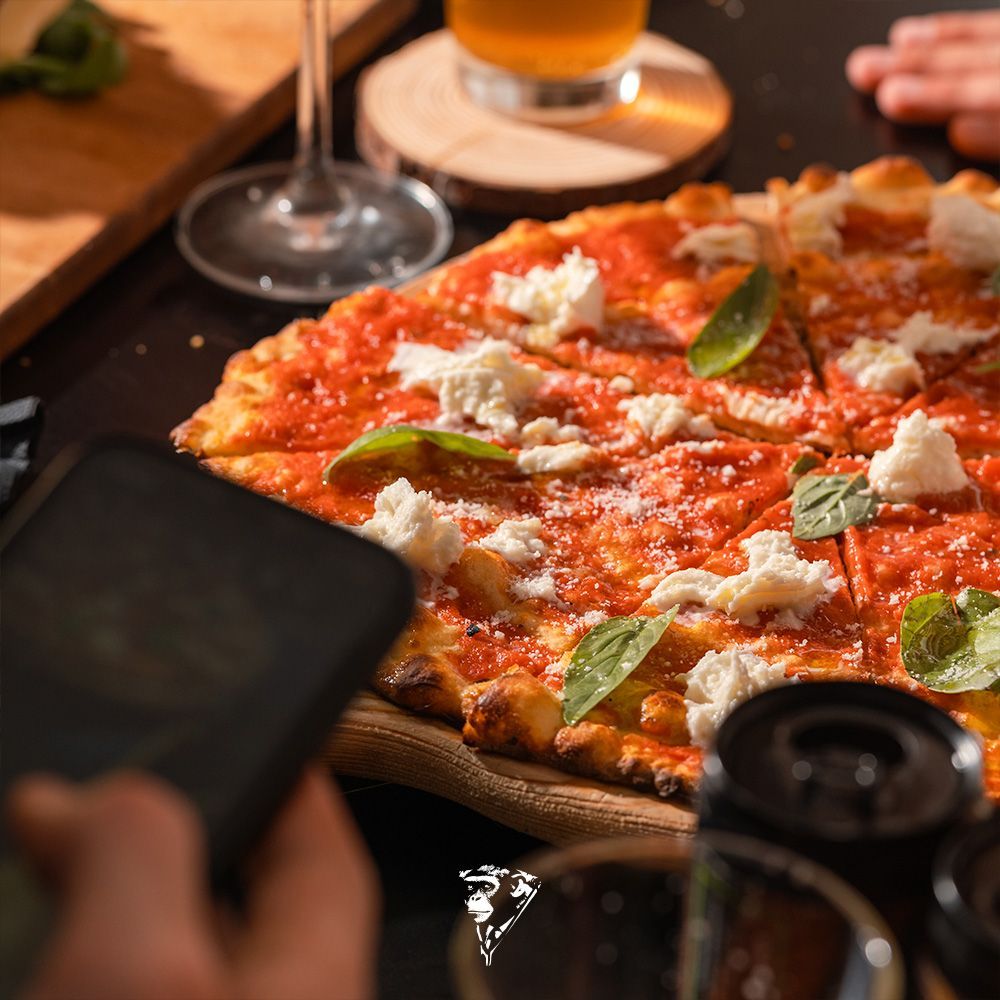 Pizza con basilico e formaggio su un tavolo, con una mano che tiene un telefono in primo piano.
