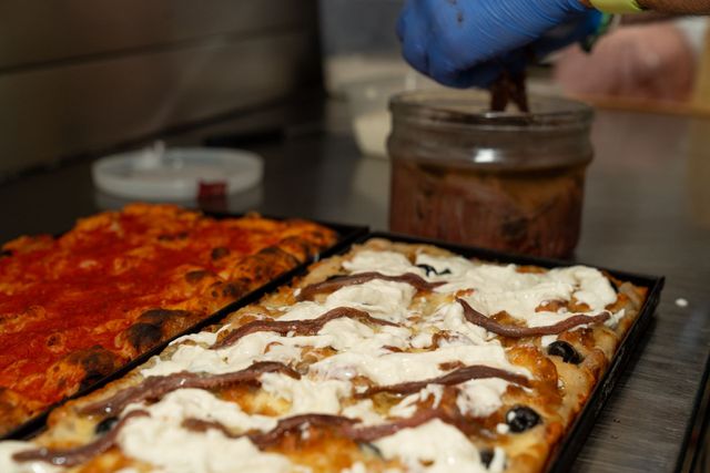 Pizza rettangolare su un tavolo, con una mano guantata e un barattolo sullo sfondo