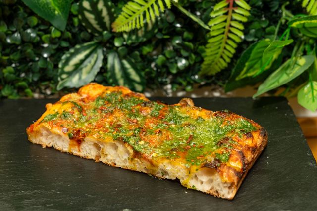 Fetta rettangolare di focaccia alle erbe su una lastra di ardesia nera, con uno sfondo di fogliame verde