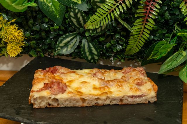 Fetta di pizza al prosciutto e formaggio su una lastra di ardesia scura, con uno sfondo di fogliame verde