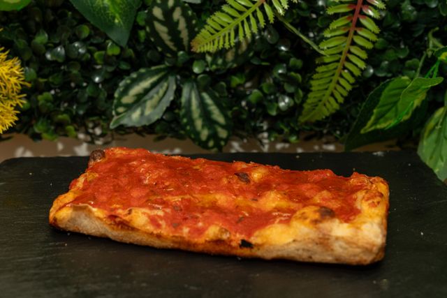 Fetta rettangolare di pizza filante su un piatto scuro, con del fogliame verde sullo sfondo