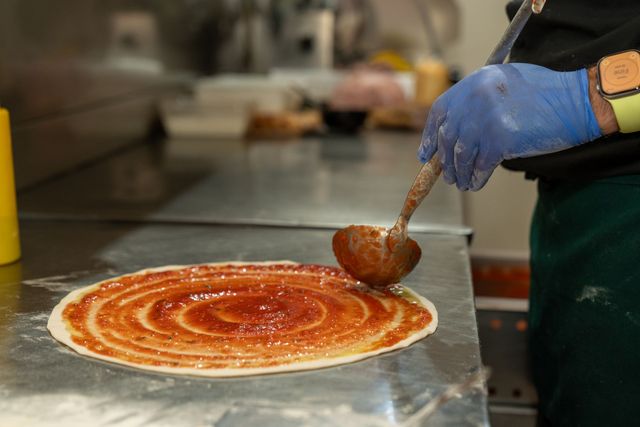 Un lavoratore con i guanti spalma la salsa di pomodoro sull’impasto della pizza su un piano di lavoro in cucina.
