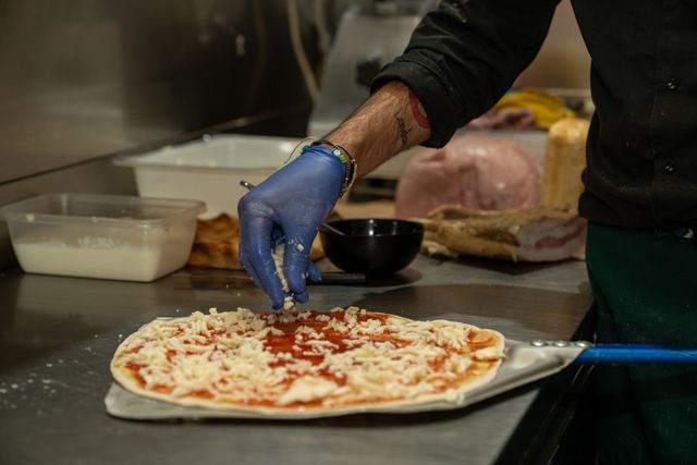 Una mano guantata spalma la salsa di pomodoro sulla base di una pizza in un’area di preparazione in cucina.