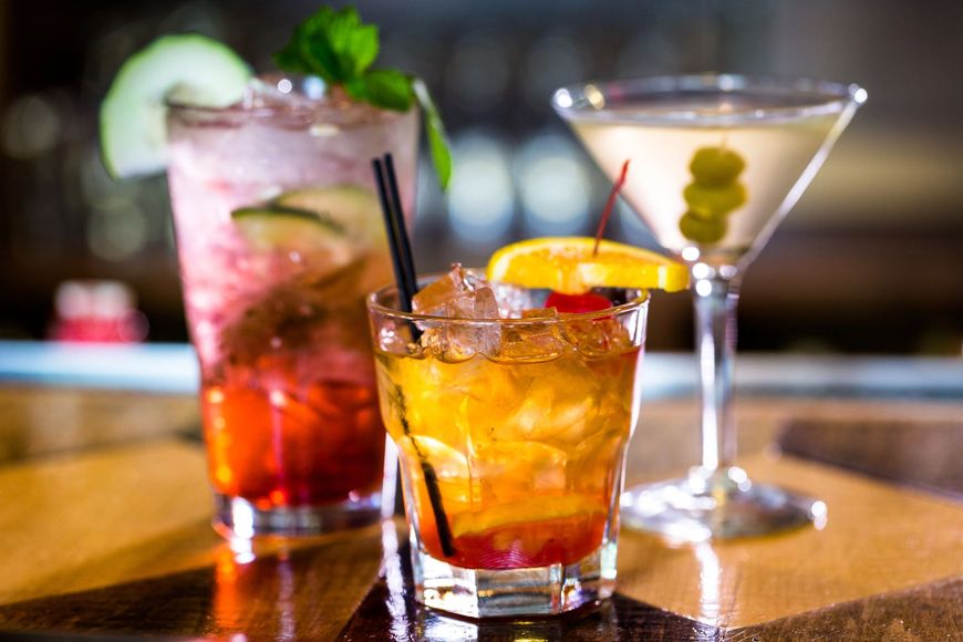 Tre cocktail colorati sul bancone di un bar, tra cui un martini con olive e bevande ghiacciate guarnite con frutta