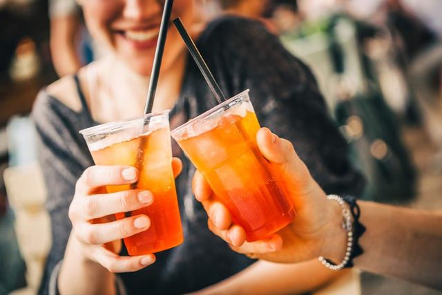 Due mani che brindano con cocktail all'arancia usando cannucce in un'ambientazione da bar sfocata.