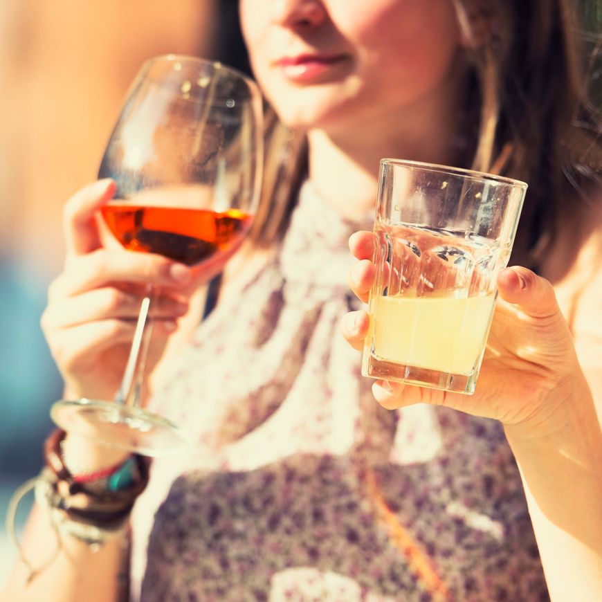 Persona che tiene in mano un bicchiere di vino rosso e un bicchiere di bevanda chiara sotto la calda luce del sole
