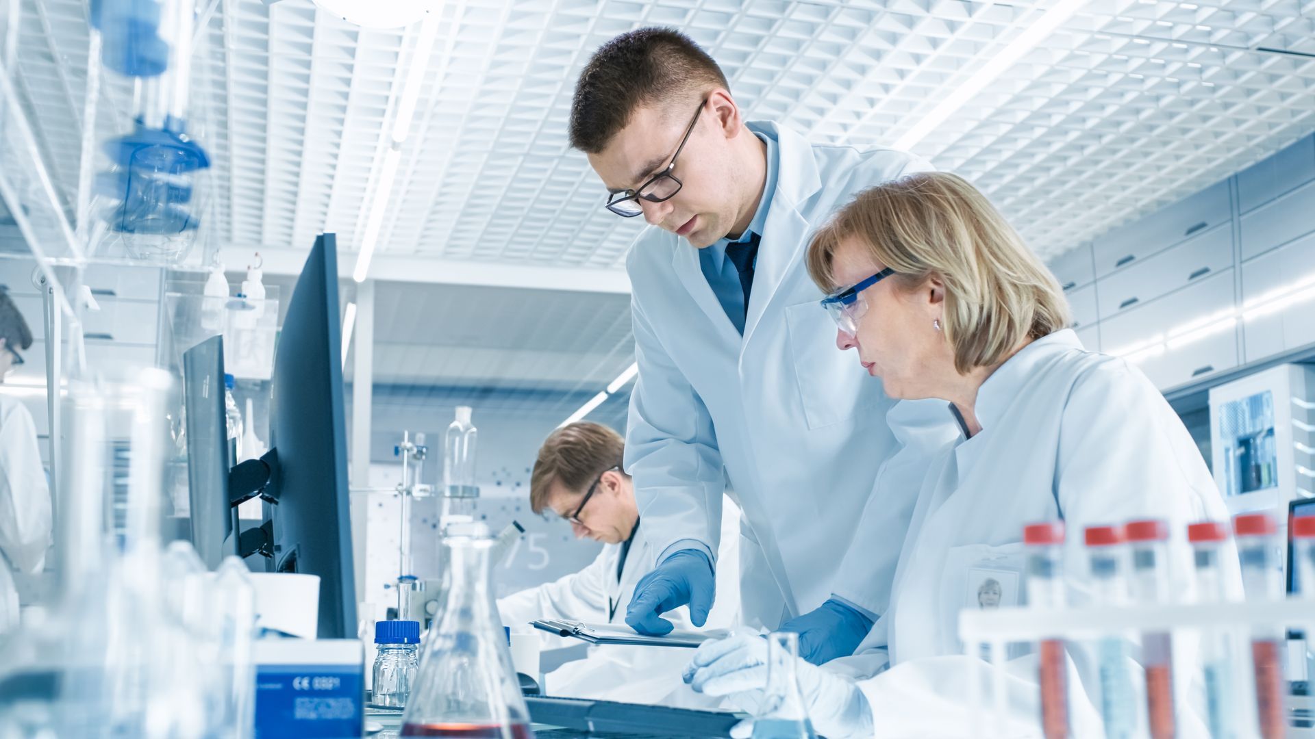 3 scientists in white coats and blue gloves working in a lab.