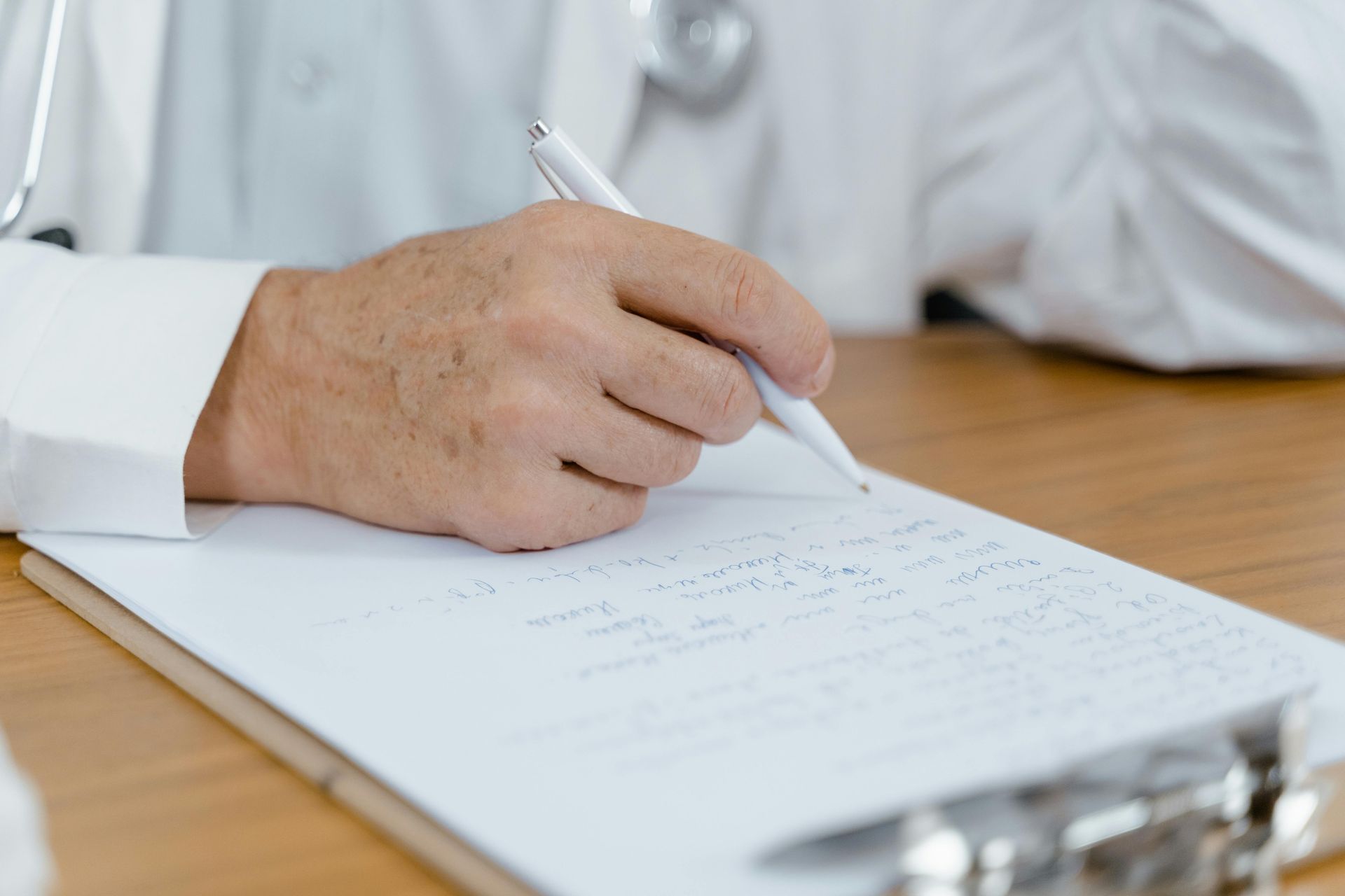 A Caucasian right hand holding a pen rests on a clipboard with writing on it. The writer wears a white lab coat.