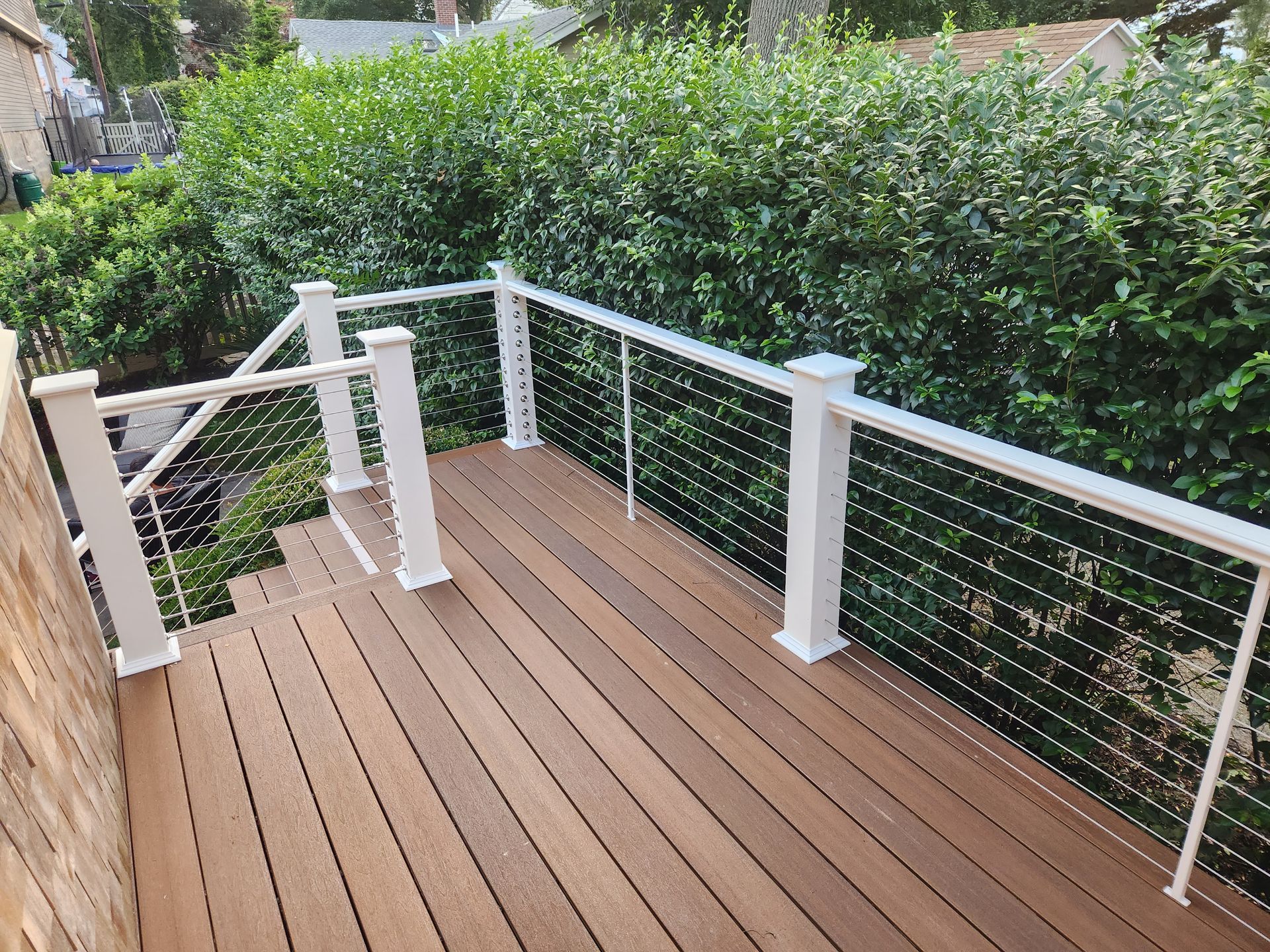 Wooden deck with white cable railing overlooking dense green hedges and a garden walkway