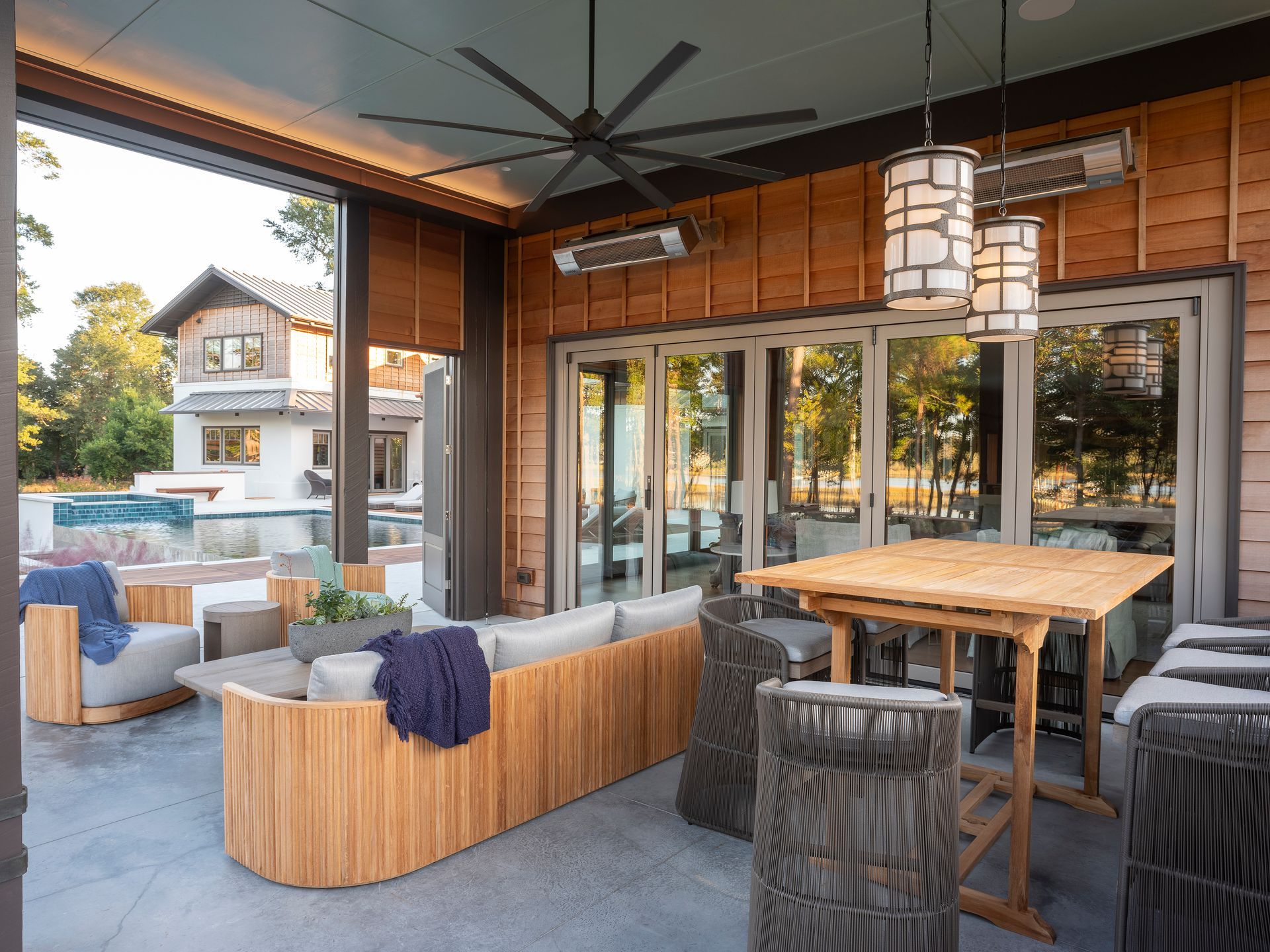 Covered outdoor patio with lounge seating, wooden dining table, and glass doors overlooking a poolside view