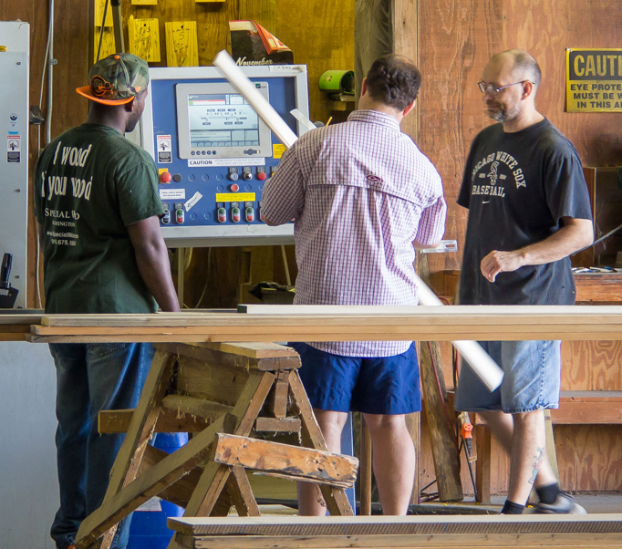 Three people standing by a wood-cutting machine in a workshop, inspecting a long white board