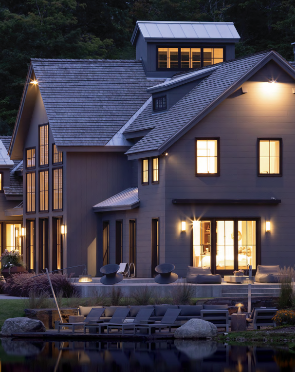 Modern gray house at dusk with warm lit windows, dockside patio, and lakeside reflections