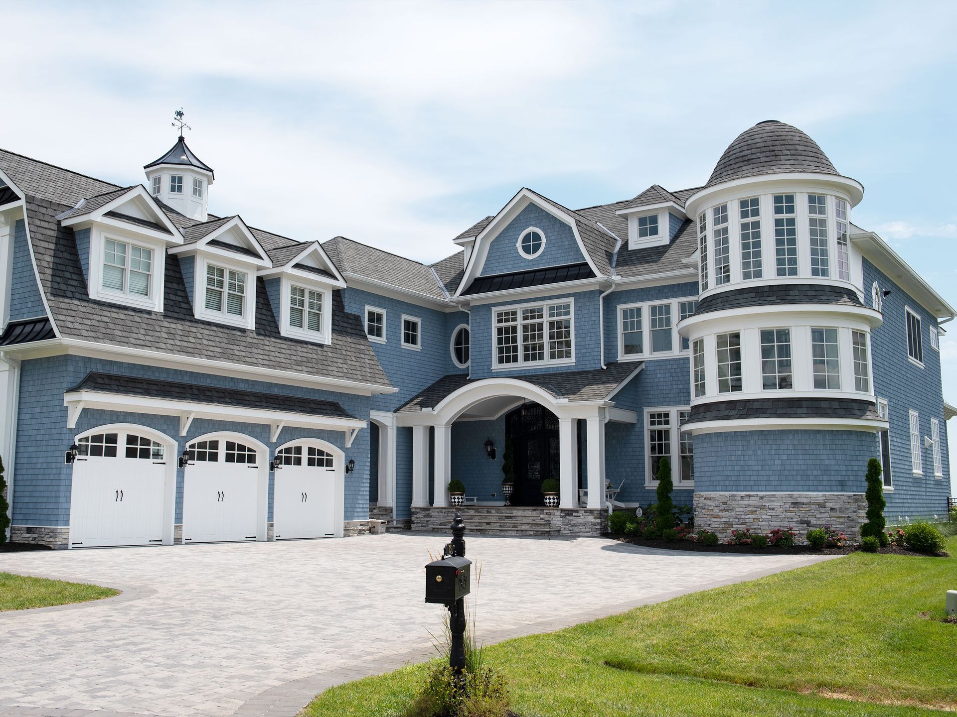 Large gray-blue suburban house with white trim, arched entryway, and a three-car garage on a landscaped lawn.