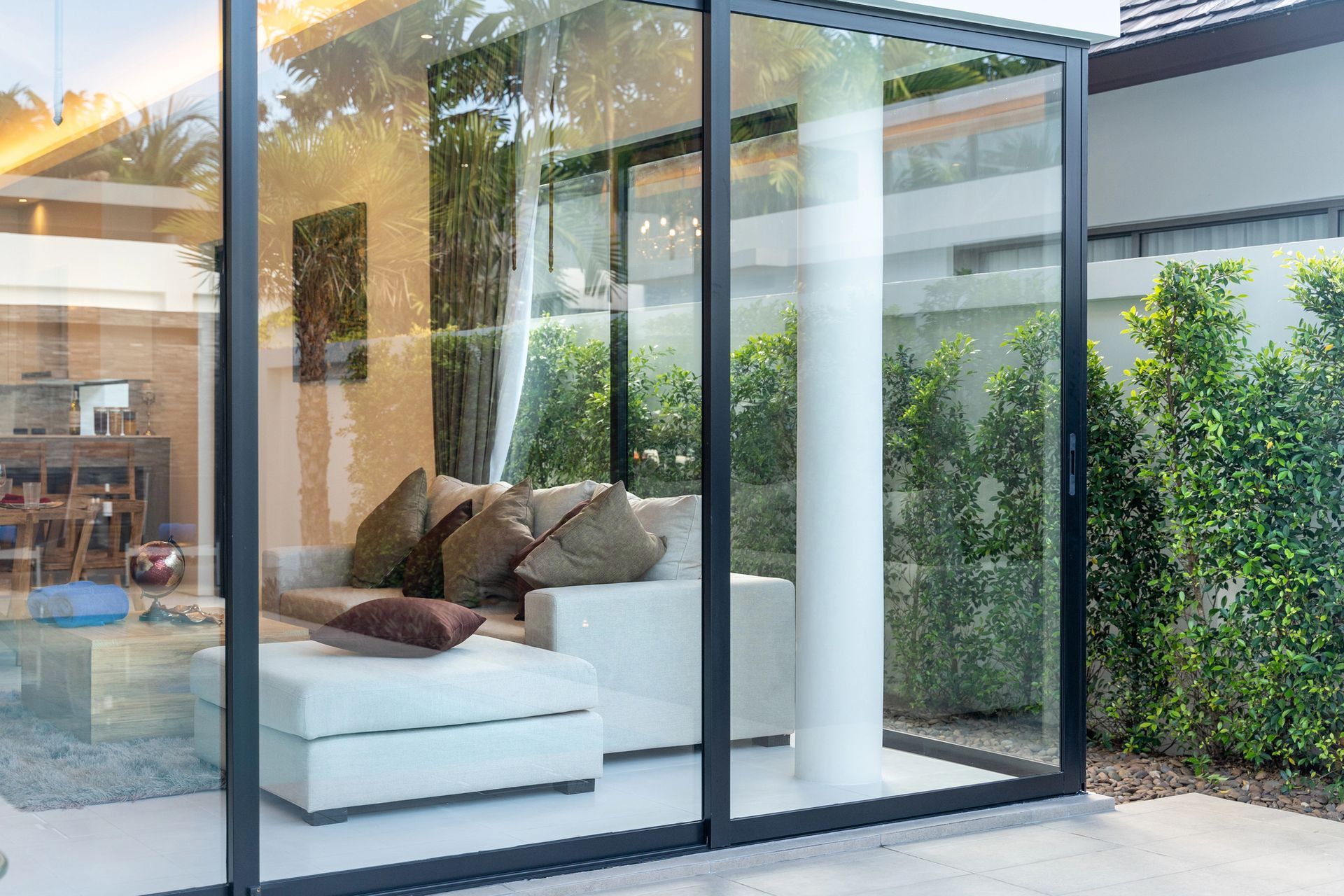 Moderno patio in vetro con divano bianco e cuscini marroni accanto a un luminoso giardino interno-esterno.