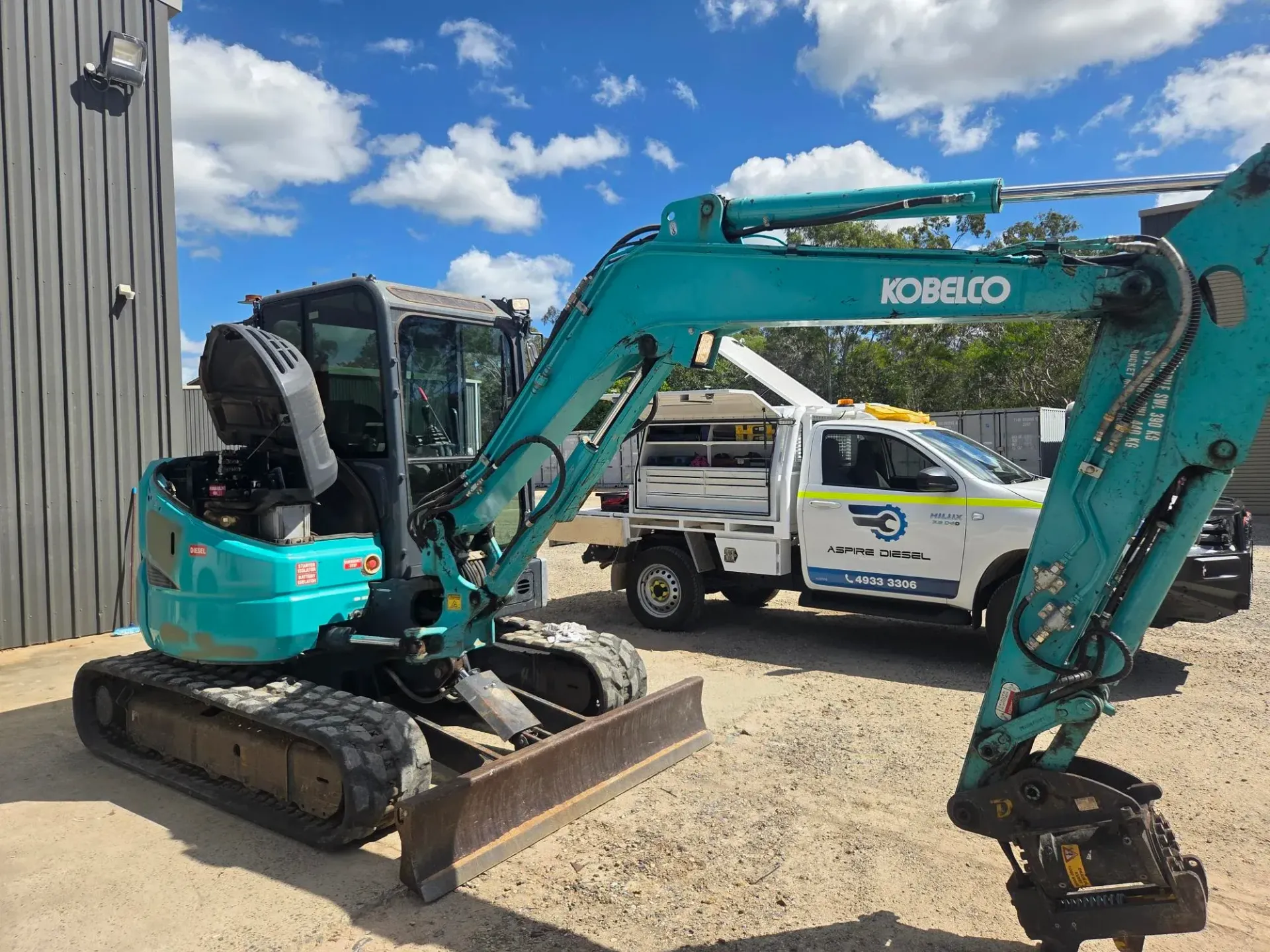 Turquoise Kobelco excavator parked beside a building on a gravel lot, with a service truck nearby — Aspire Diesel In Gracemere, QLD