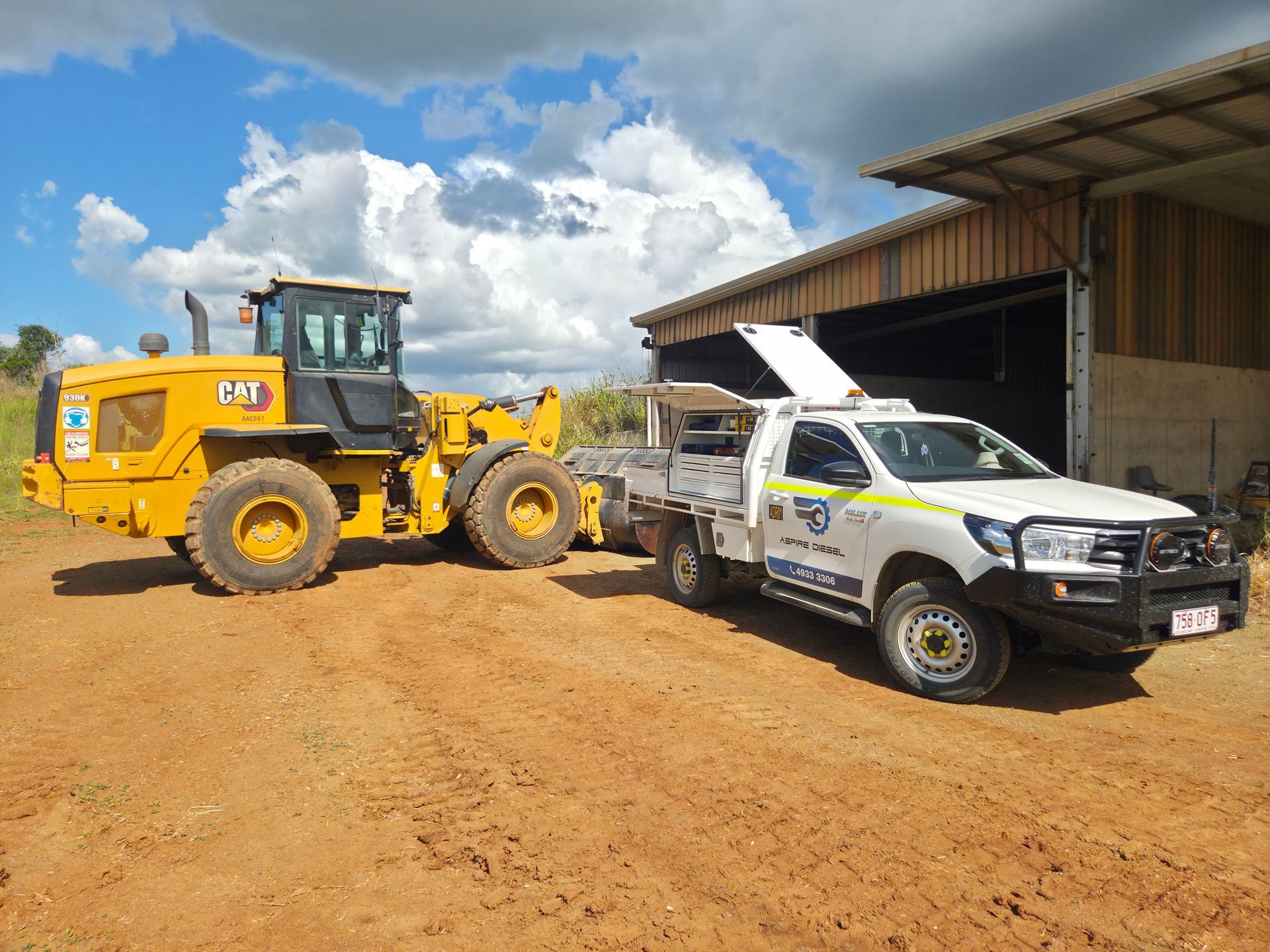 Yellow front loader beside a white utility truck outside a barn on a dirt lot under a cloudy sky — Aspire Diesel In Gracemere, QLD