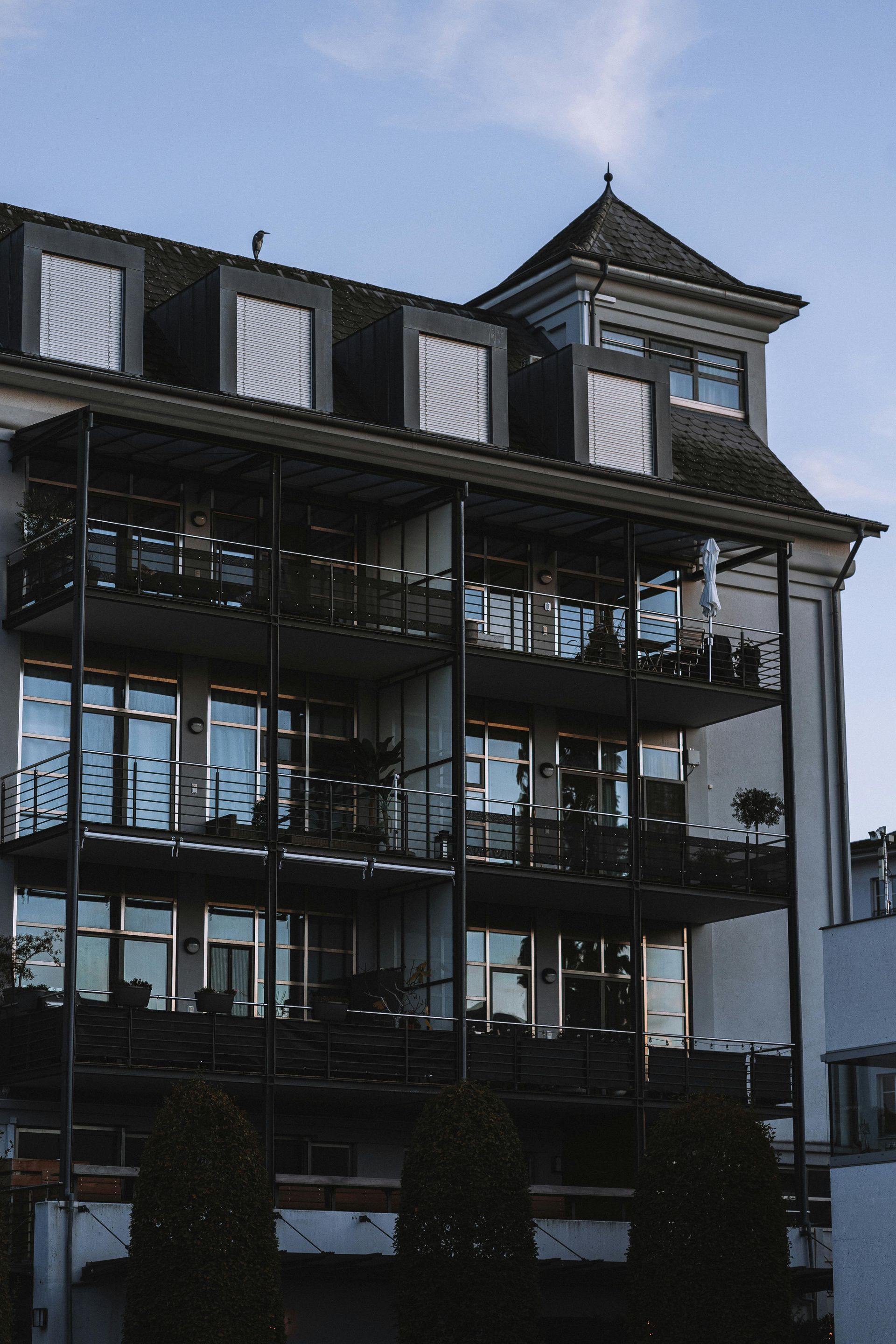 A white apartment building with tiered balconies, metal railings, and a small tower on the roof under a blue sky.