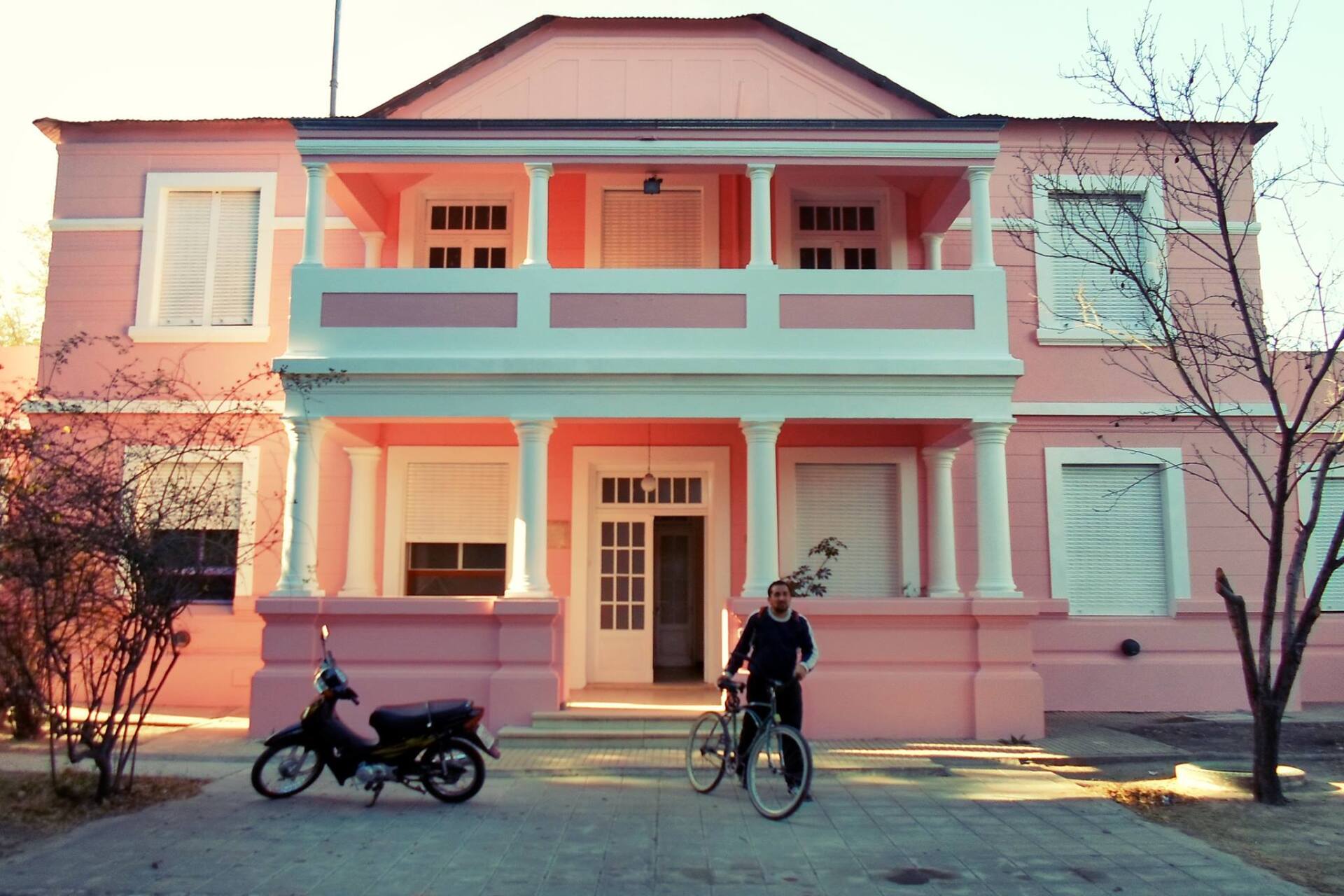 Edificio rosa de dos pisos con balcón, una persona con una bicicleta y una motocicleta estacionada.