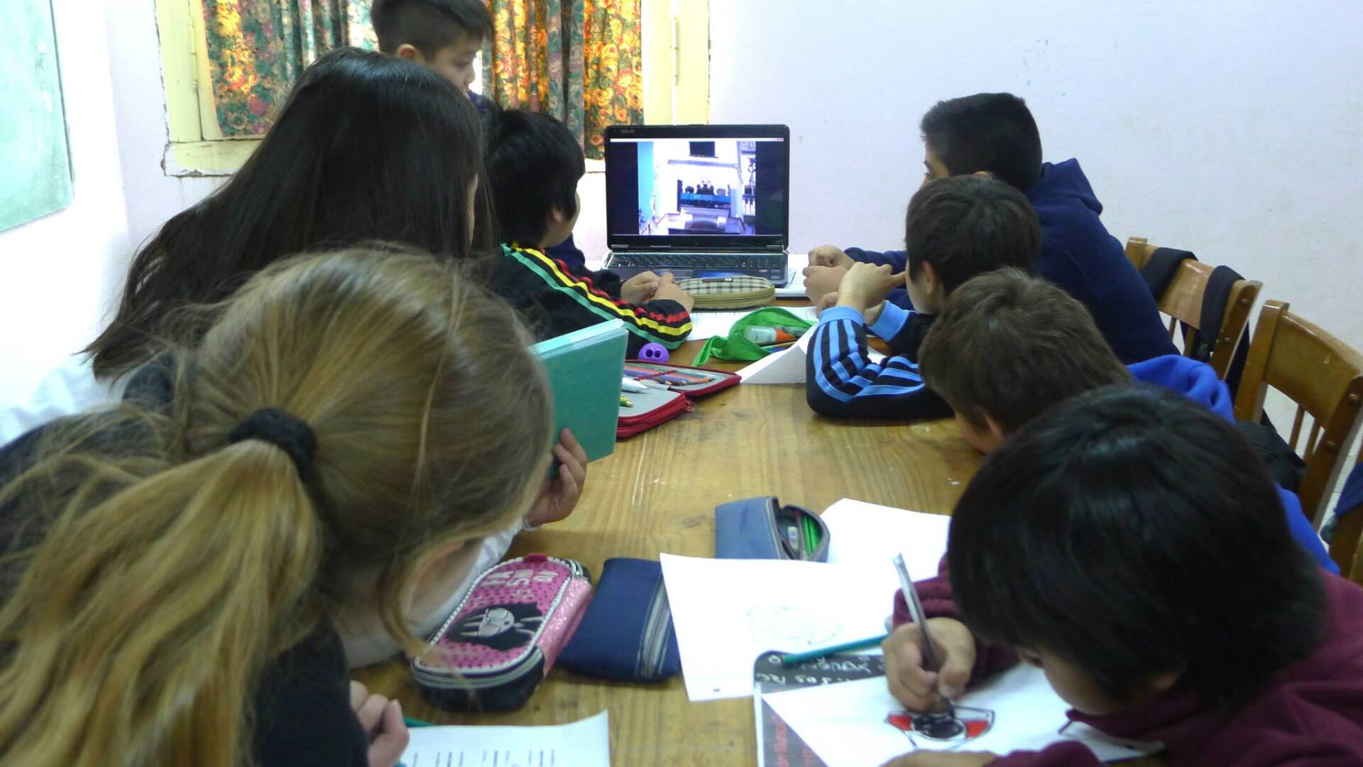Niños reunidos alrededor de una mesa, mirando la pantalla de una computadora portátil. Algunos escriben en papel. Habitación luminosa.