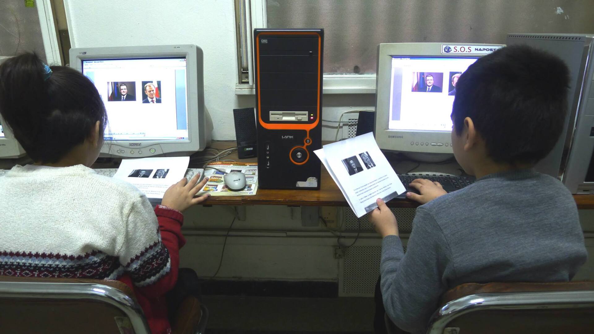 Dos niños frente a computadoras, mirando pantallas y papeles; computadoras e imágenes impresas en un escritorio.