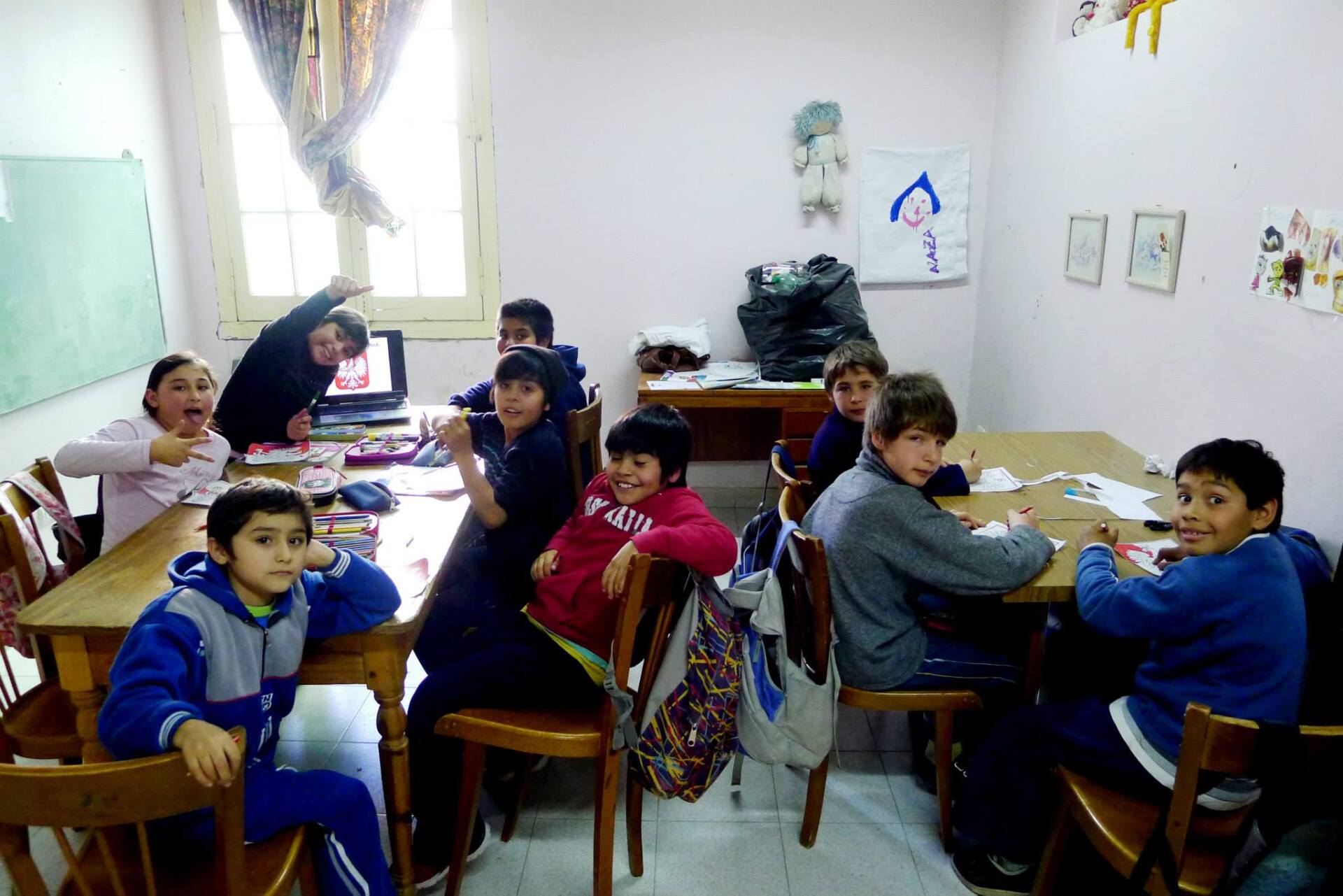 Niños en un aula, algunos sentados en mesas dibujando, otros mirando a la cámara.