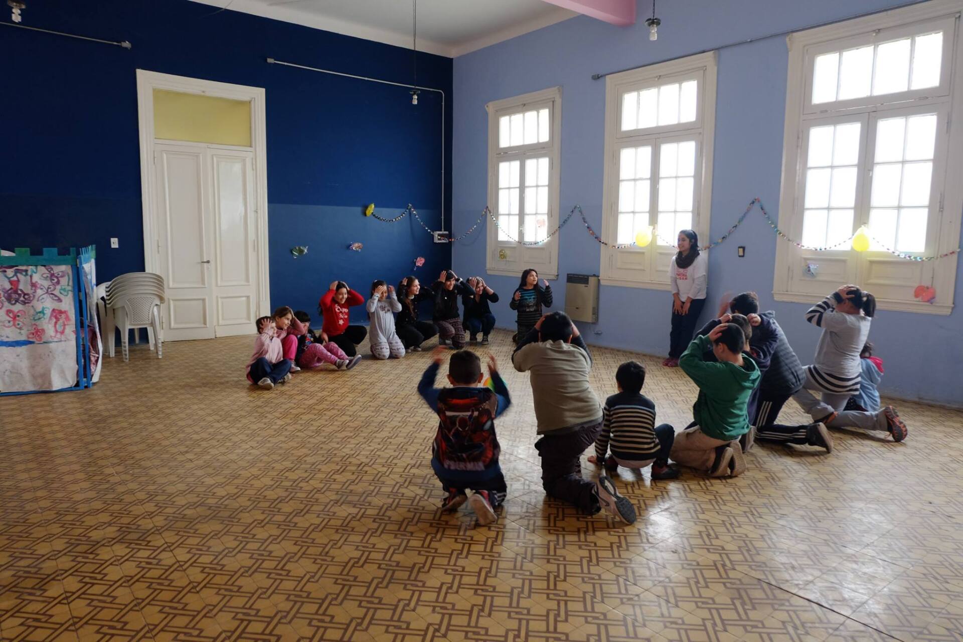 Niños y adultos en una habitación, algunos arrodillados, todos señalando con las manos como si estuvieran tomando fotografías.