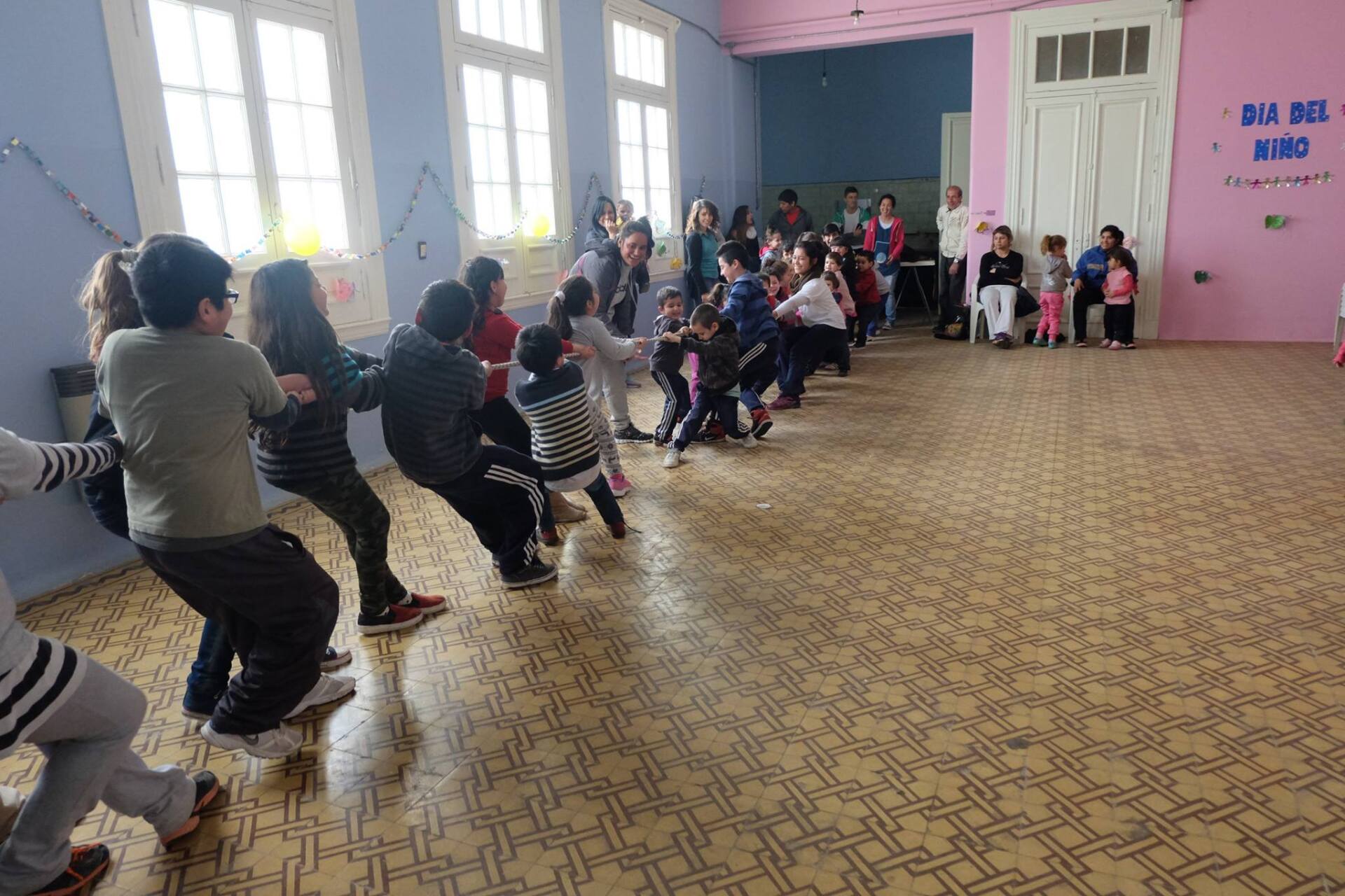 Niños jugando al tira y afloja en un espacio cerrado, tirando de una cuerda. Los espectadores observan en una sala colorida.