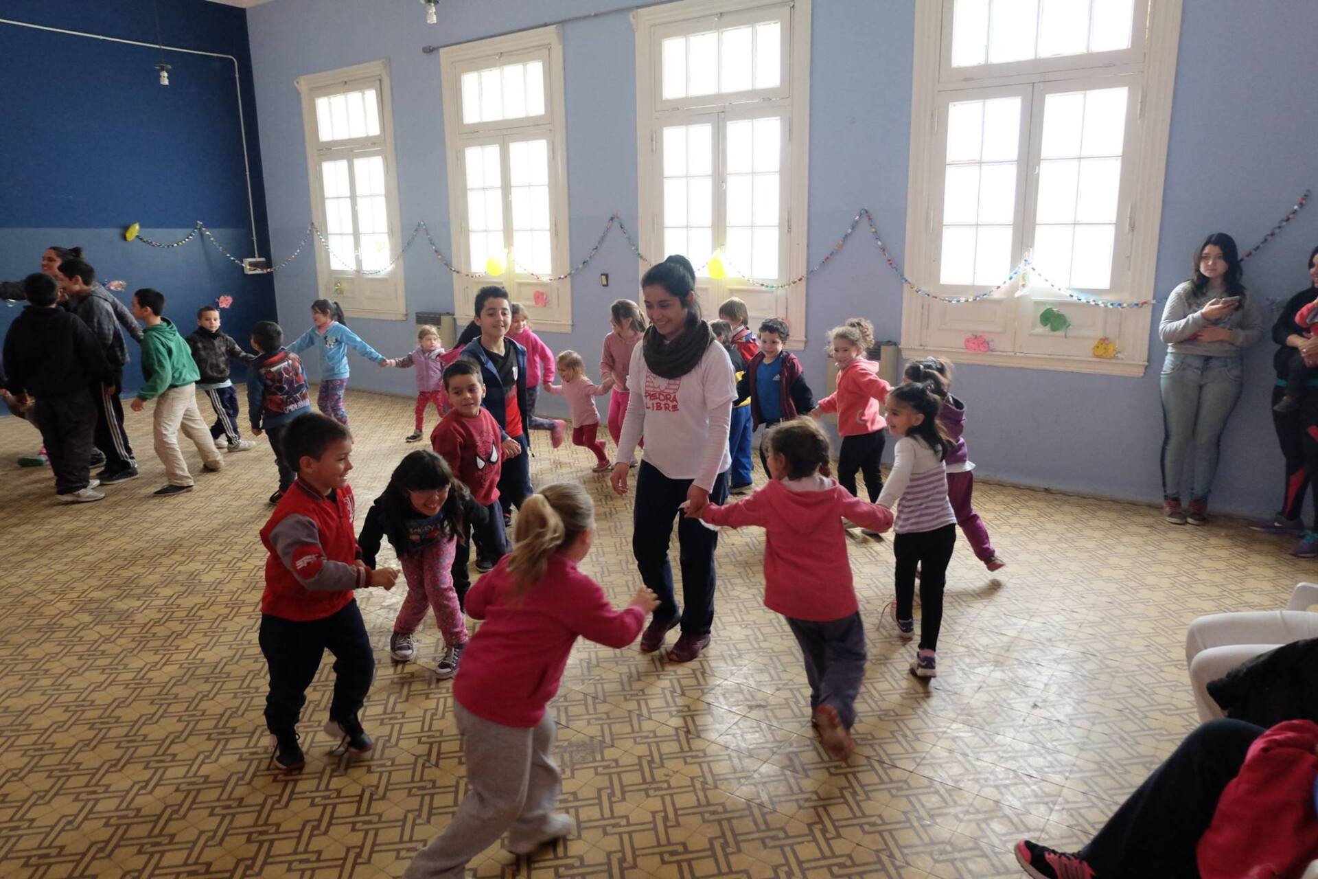 Niños tomados de la mano en círculo, guiados por una mujer, en una habitación con ventanas.