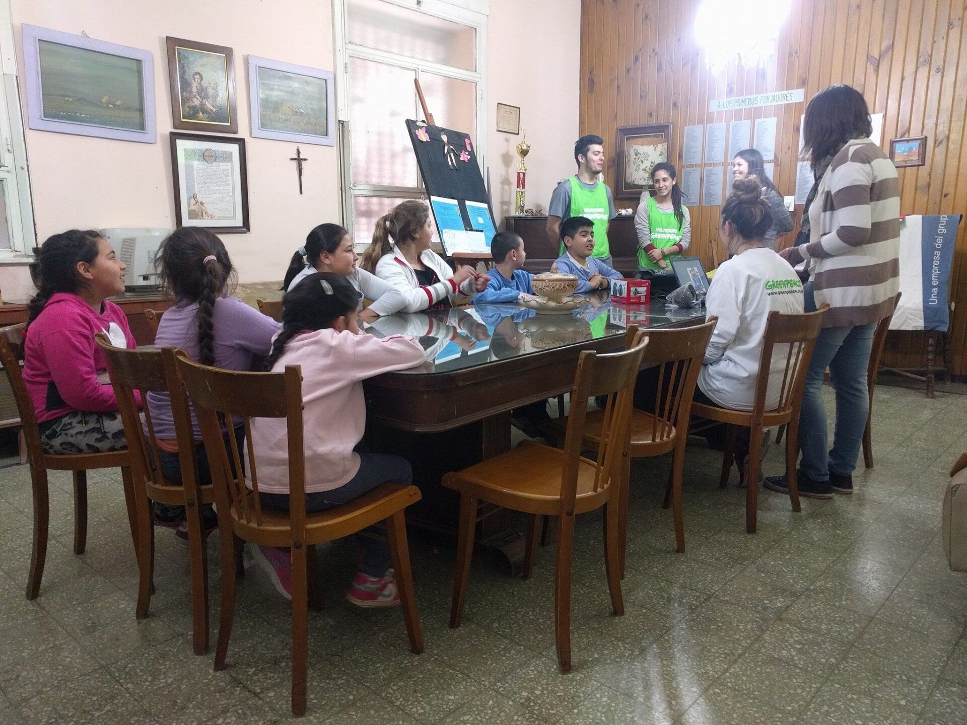 Niños y adultos en una sala reunidos alrededor de una mesa; se está realizando una presentación.