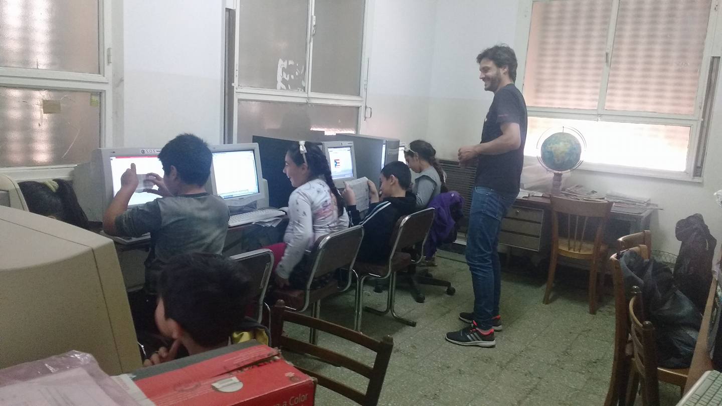 Niños y un hombre en un laboratorio de informática. Los niños están sentados frente a las computadoras, y el hombre, de pie junto a los escritorios, sonríe.