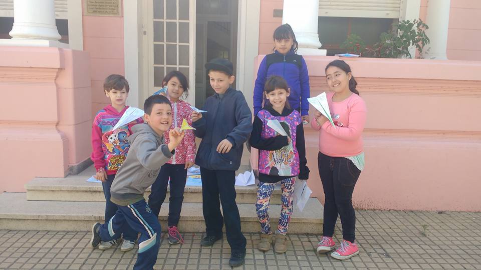 Niños sosteniendo aviones de papel posan en los escalones frente a un edificio rosa.
