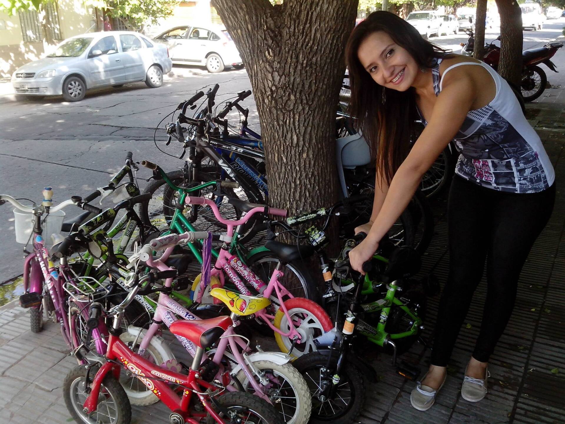 Mujer sonriendo junto a una pila de coloridas bicicletas infantiles estacionadas junto a un árbol en una calle de la ciudad.