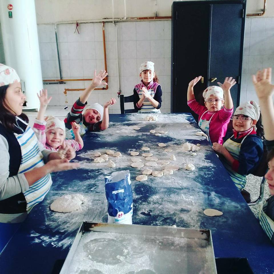 Niños horneando, saludando desde una mesa de cocina cubierta de harina y masa. Llevan delantales y gorros de chef.