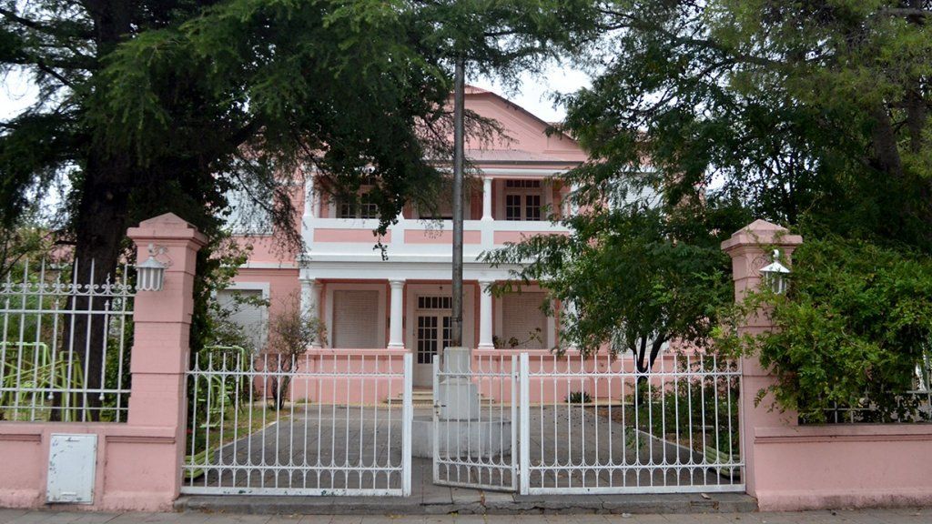 Casa rosa de dos pisos con cerca blanca y portón parcialmente abierto, árboles rodean el edificio.