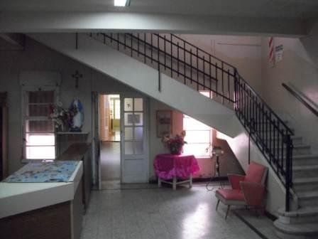 Vestíbulo interior con escalera, mostrador de recepción y estatua religiosa. Silla rosa y mesa con flores.