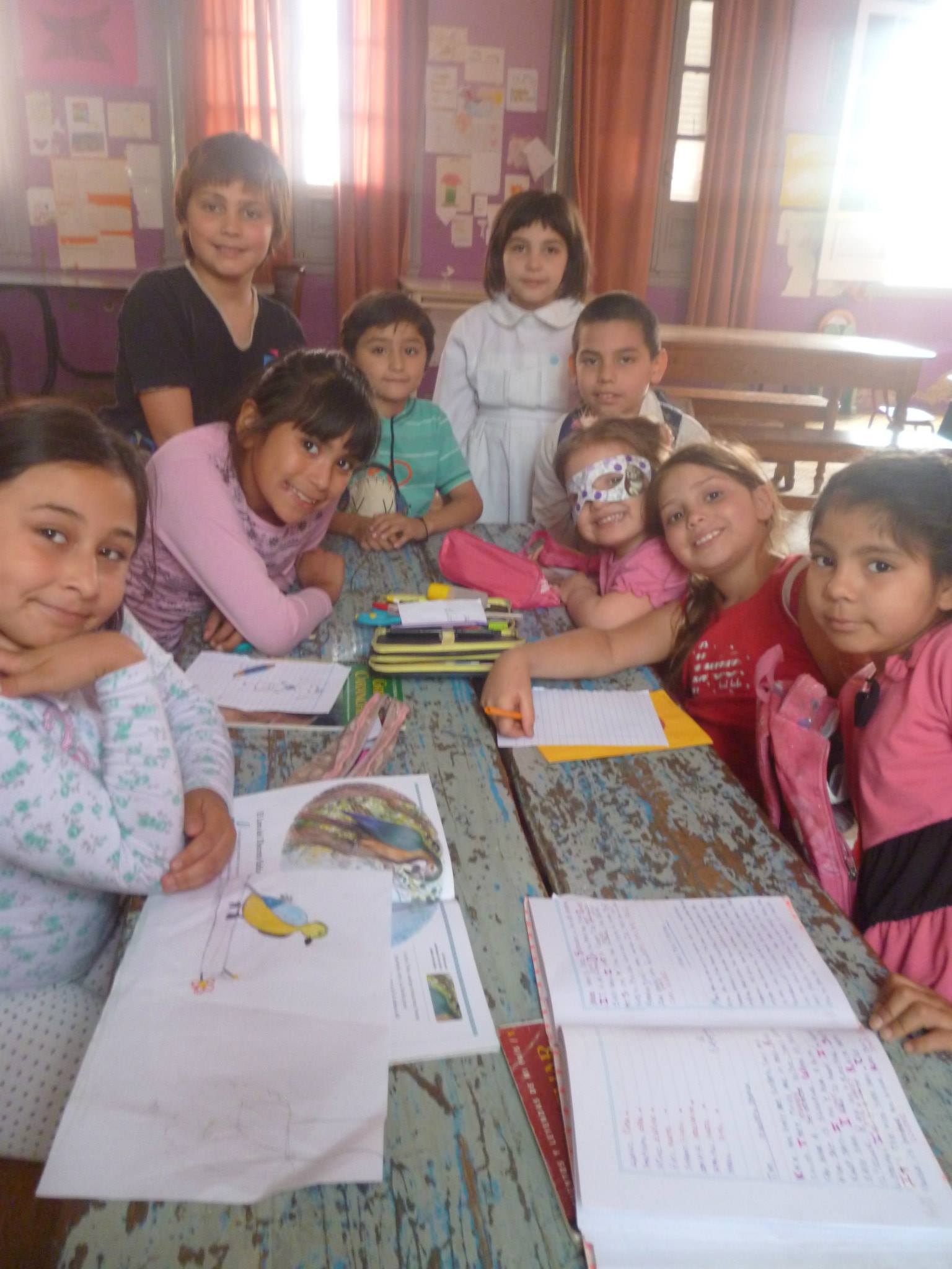 Niños en una mesa de madera en un aula; algunos mirando un libro, otros a la cámara.