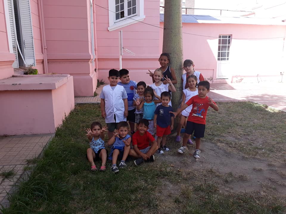 Grupo de niños posando cerca de un edificio rosa y un árbol en un día soleado.