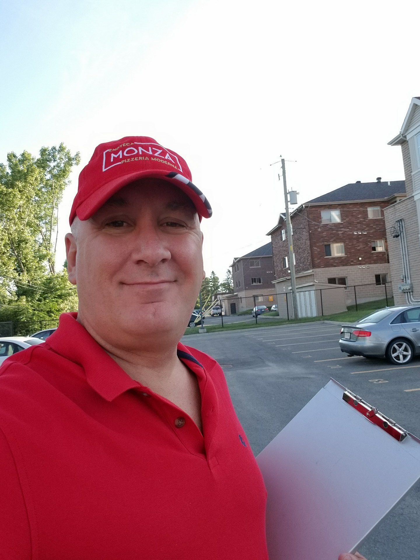 Un homme portant un chapeau et une chemise rouges, tenant un bloc-notes, sourit. À l'extérieur, près d'immeubles d'habitation.