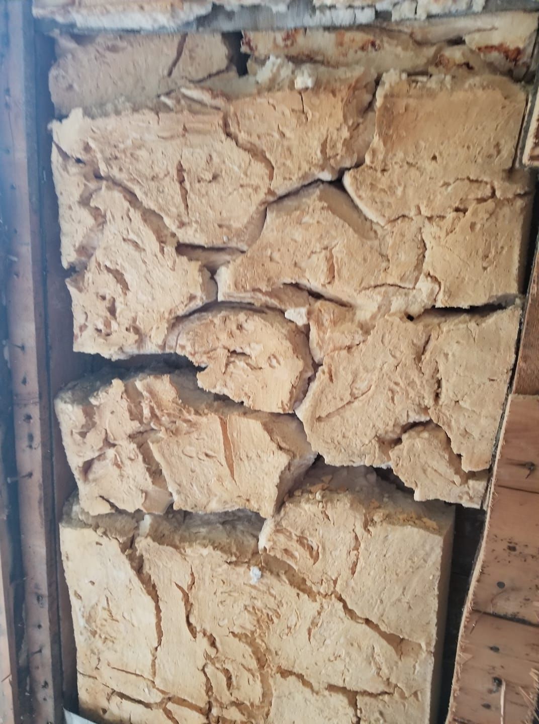 Gros plan sur un mur brun clair de construction sommaire. De grosses pierres irrégulières sont disposées en couches. On aperçoit une charpente en bois.