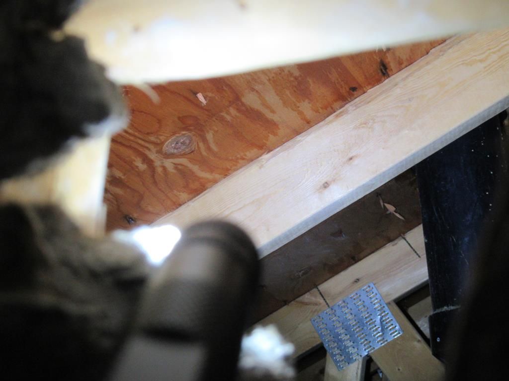 Vue depuis un grenier sombre ; charpente en bois, contreplaqué, conduits de ventilation et isolation visibles.