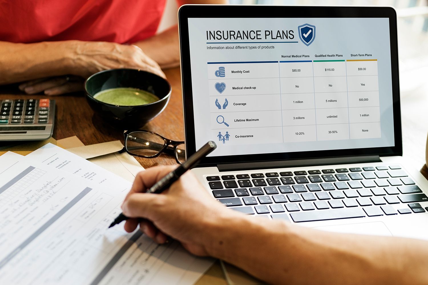 Person writing on paperwork, laptop displaying insurance plan options, calculator and bowl of liquid nearby.
