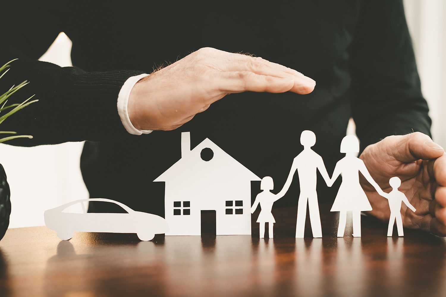 Hand protecting paper cutouts of a family, house, and car on a desk.