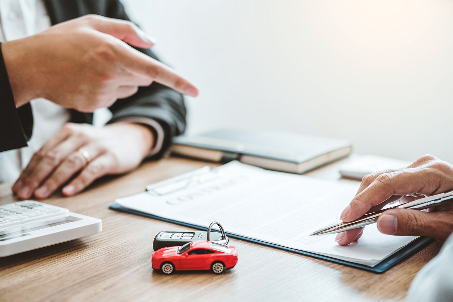 Two people at a desk; one points while the other signs a document. A toy red car and car keys sit on the desk. Two people at a desk; one points while the other signs a document. A toy red car and car keys sit on the desk.