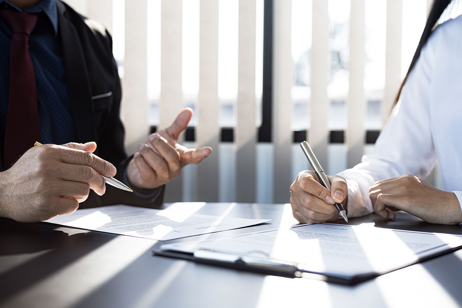 Person in suit gestures, while another signs document at table. Sunlight streams through blinds.