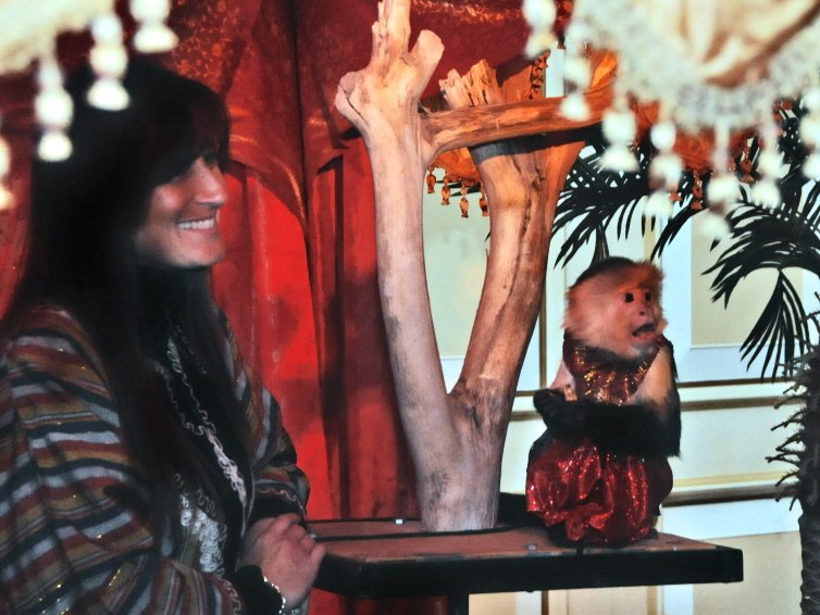 Woman smiles near a capuchin monkey wearing a red skirt, posing on a wooden stand.