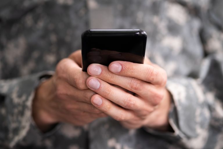 Person in camouflage uniform holding a black smartphone, typing.
